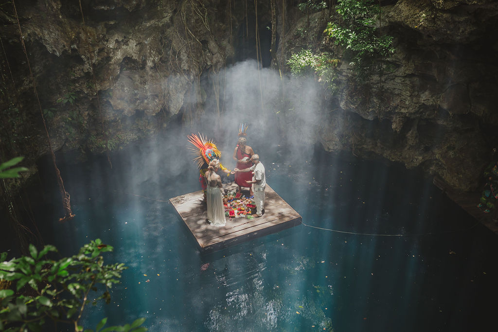 Una pareja ante el altar maya sobre una plataforma de madera en medio de un cenote, bajo un haz de luz