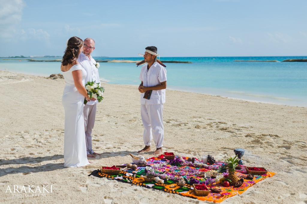 Una pareja vestida de blanco frente a un oficiante de blanco en la playa, con una ofrenda de colores sobre la arena