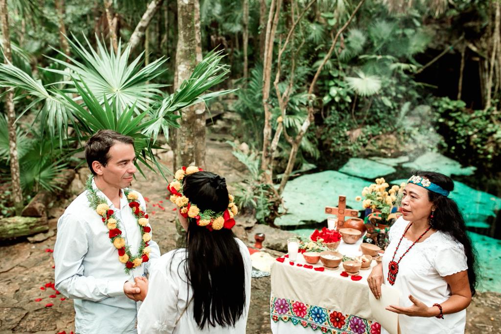 Una pareja con collares de flores ante una guía maya de blanco junto al altar, en la selva